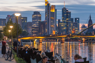 Frankfurt skyline at dusk viewed from the banks of the Main River, with illuminated skyscrapers, city lights reflecting on the water, and people walking and sitting along the riverside promenade.