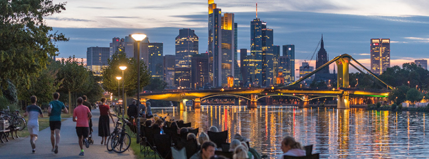 Frankfurt skyline at dusk viewed from the banks of the Main River, with illuminated skyscrapers, city lights reflecting on the water, and people walking and sitting along the riverside promenade.