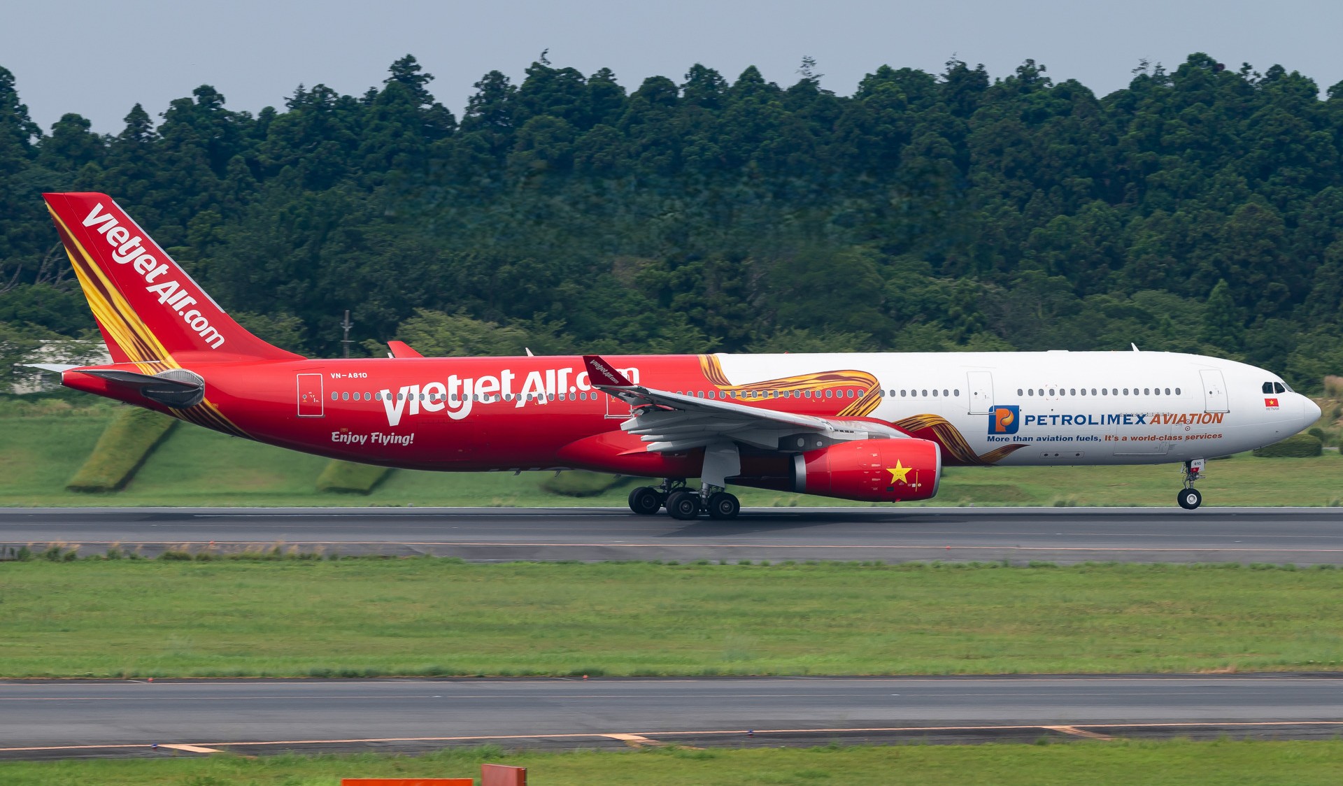 Red and white Vietjet Airbus A330-300, registration VN-A810, on a runway with a dense green tree line behind it.